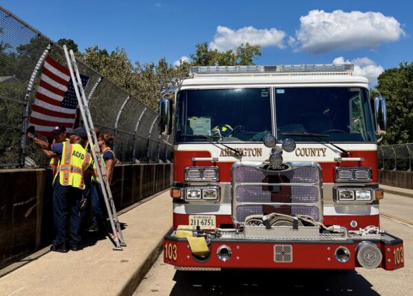 Firefighters in high-visibility vests set up an American flag along a chain-link fence beside a red Arlington County fire engine.