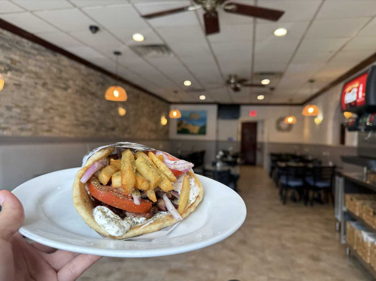 Loaded pita sandwich with fries, tomato slices and red onions on a white plate, held in a casual restaurant.