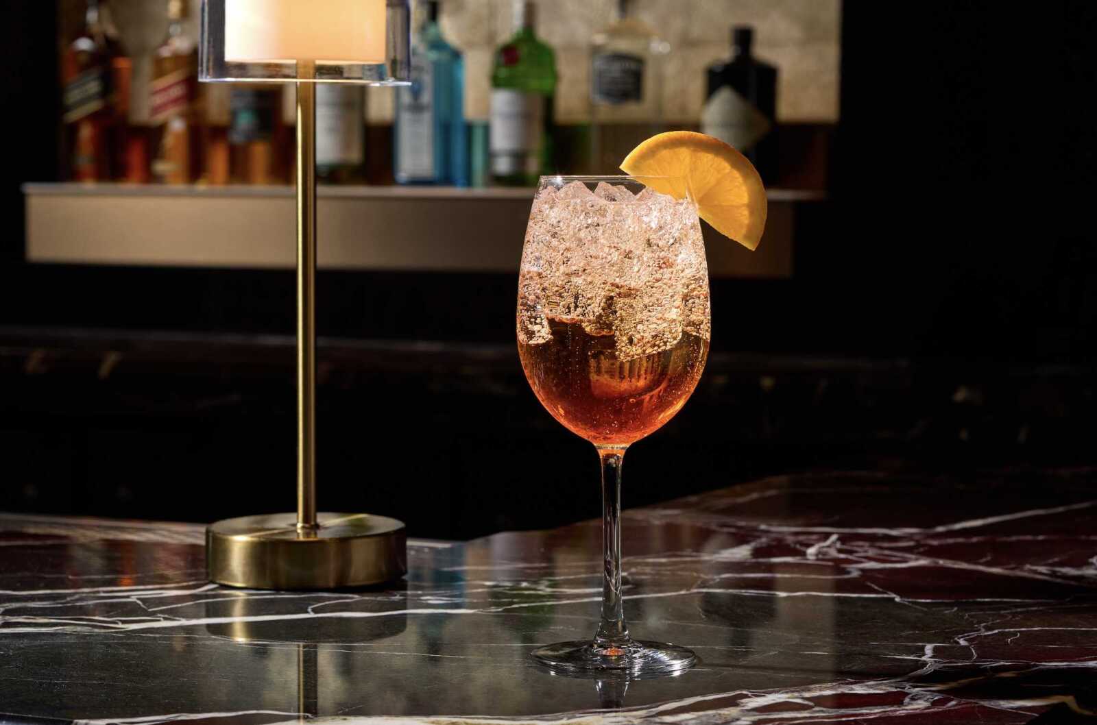 Cocktail glass with ice and an orange slice on a marble bar, dimly lit behind a row of bottles.