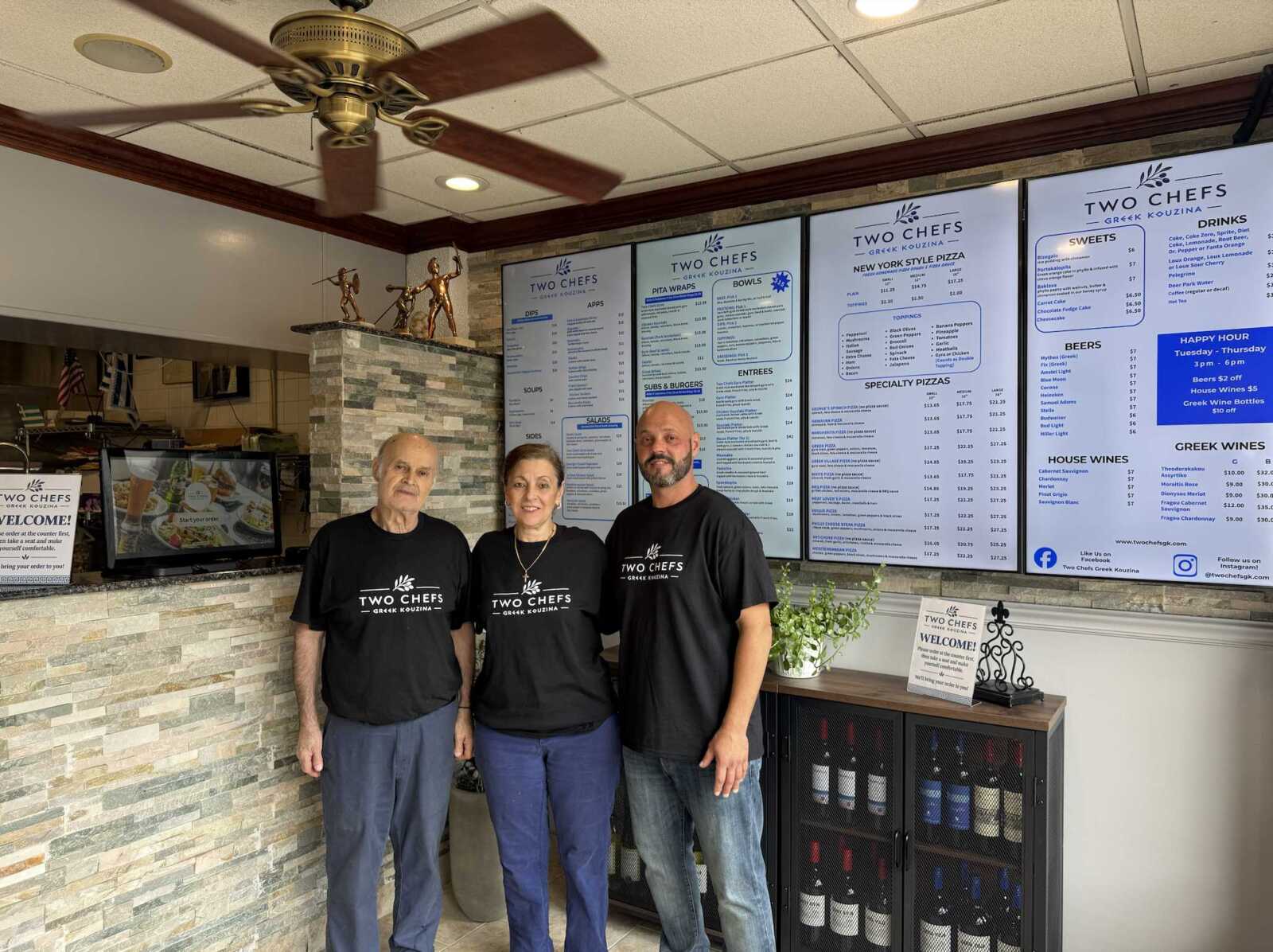 Three staff members in black Two Chefs shirts stand in front of a counter with large menu boards on the wall inside a Greek restaurant.