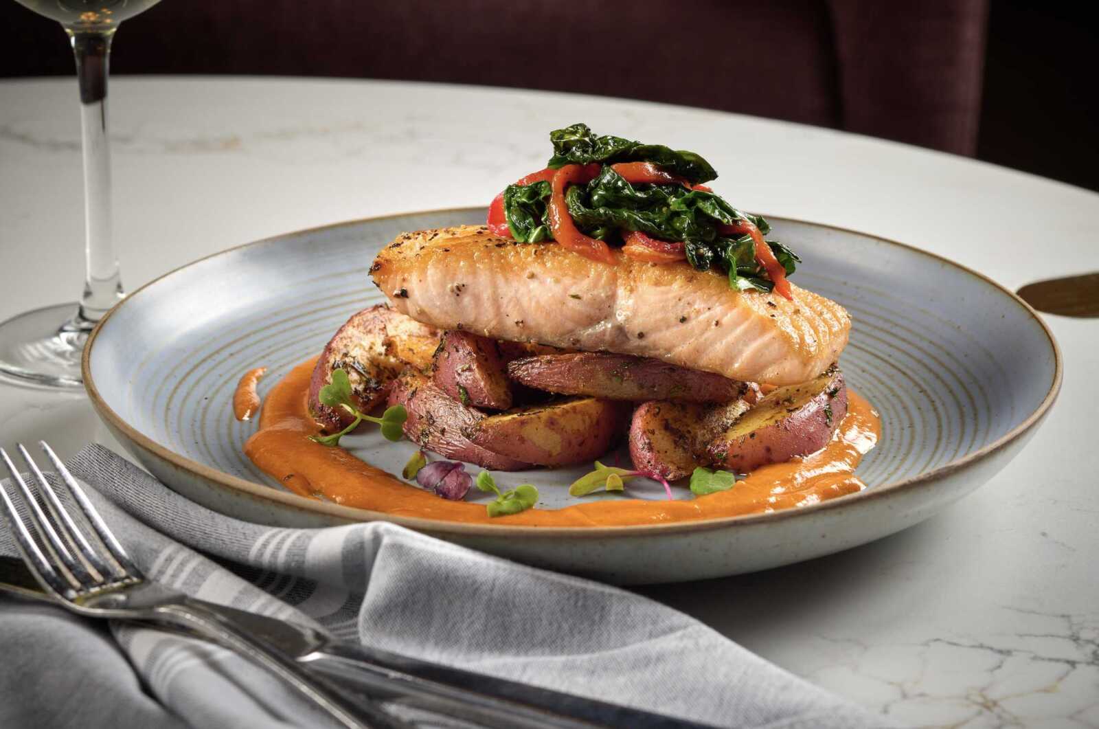 Salmon fillet perched on roasted potato slices with orange sauce and sautéed greens on top of a blue plate, wine glass in the background.