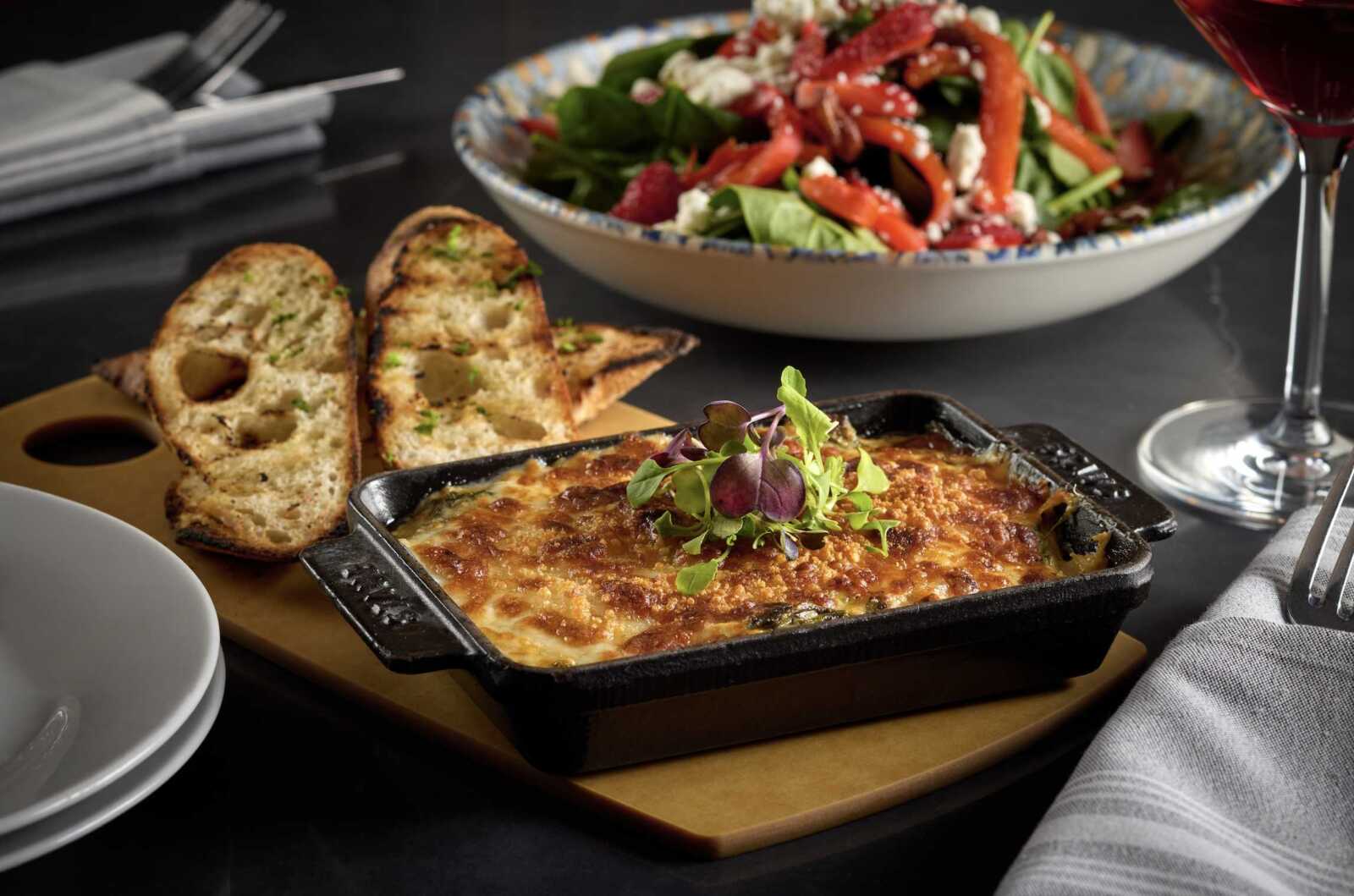 Baked cheesy casserole in a cast‑iron dish topped with greens, with sliced bread and a salad in the background on a dark table.