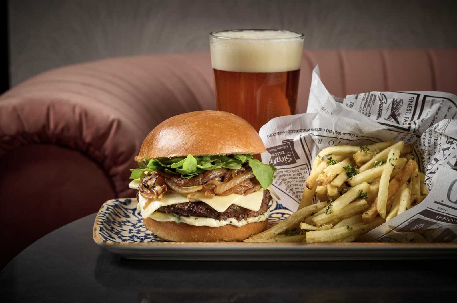Juicy burger with lettuce, onions, cheese, and sauce on a glossy bun beside a stack of seasoned fries on a newspaper-wrapped tray, plus a beer behind.