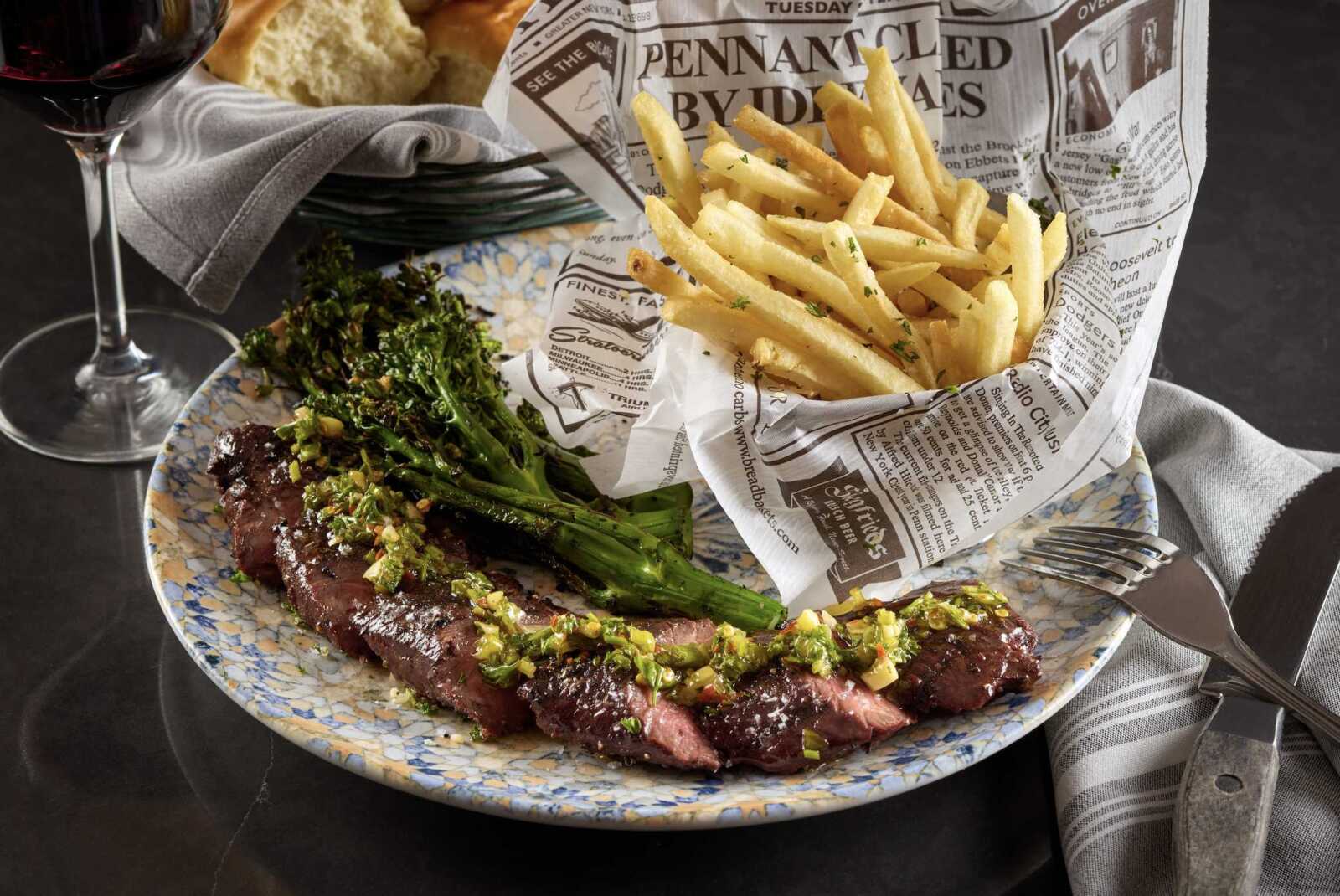 Plate of sliced steak with a chopped herb garnish served with broccoli rabe and a heap of French fries wrapped in newspaper, plus a glass of red wine in the background.