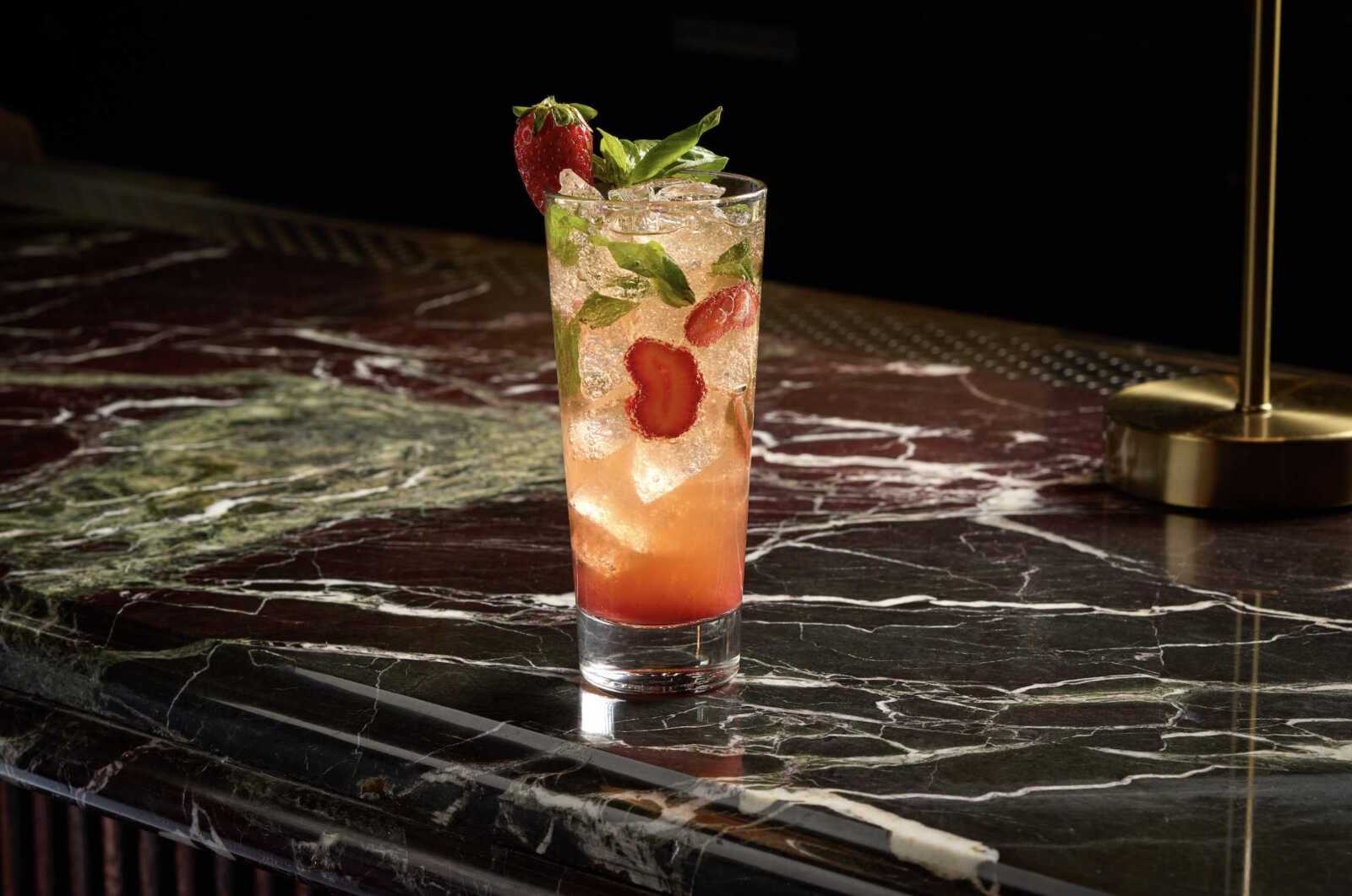 Tall cocktail glass with strawberry and mint garnish on a dark marble bar, ice-filled pink drink.