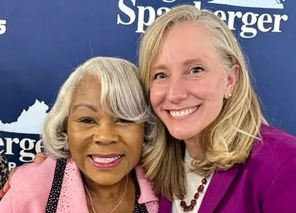 Two women smiling for a photo, one older with gray hair in a pink cardigan and the other a younger blonde in a magenta jacket, standing before a blue backdrop with white text.