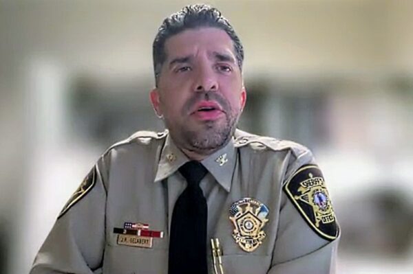 Male police officer in a tan uniform with a black tie speaking, badge visible on chest.
