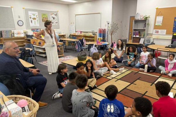 Teacher stands with hands on hips addressing a diverse circle of elementary students seated on a colorful rug in a classroom.