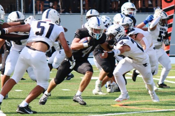 Running back in black jersey carries the ball as white-uniform defenders close in on a crowded line.