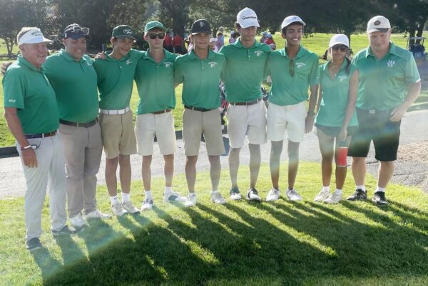 Group of golfers in green polo shirts posing together on a sunny course, arms around each other.