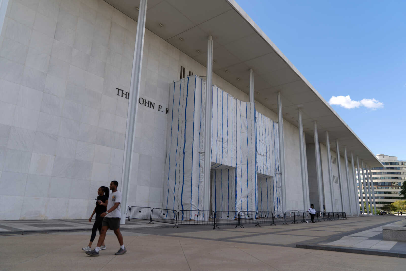 White marble building with tall columns and a blue-striped wrapped entrance, with pedestrians walking by and temporary barriers in front.