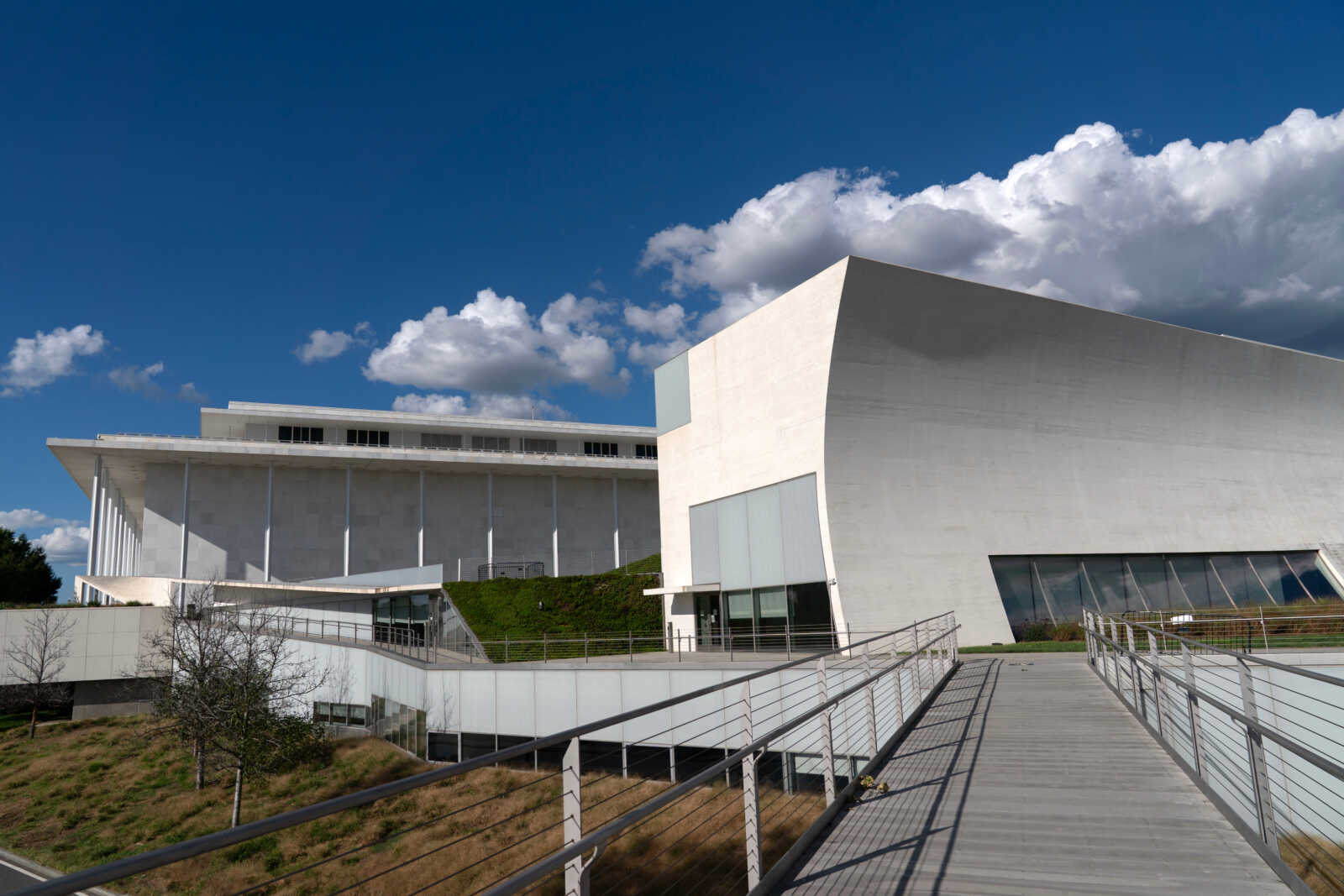 Modern white concrete building with a walkway and metal railing leading toward glass doors under a blue sky with clouds, on a sunny day.