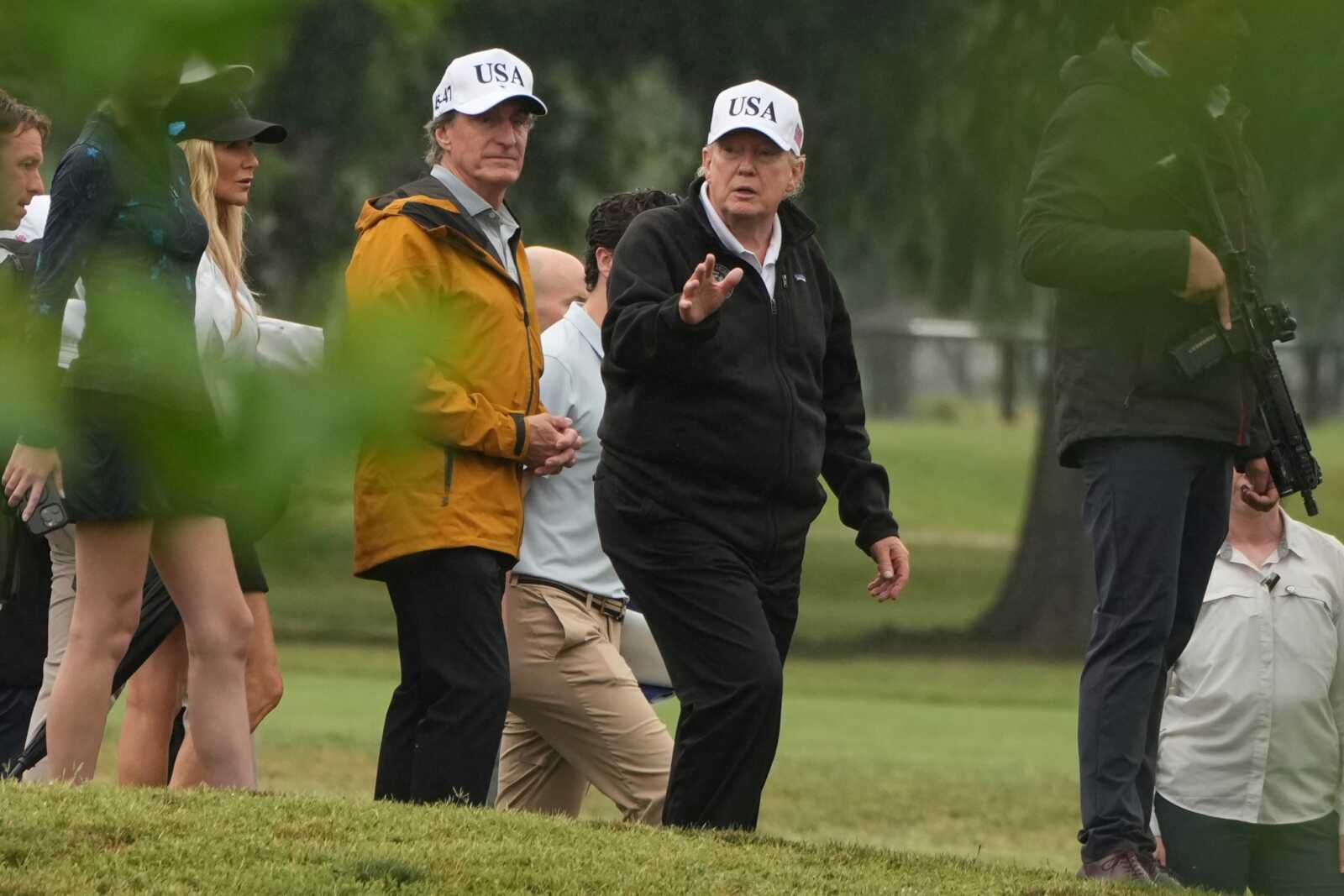 Group of people walking on a grassy area; two men wear white USA caps, one in a black jacket gestures as others follow.