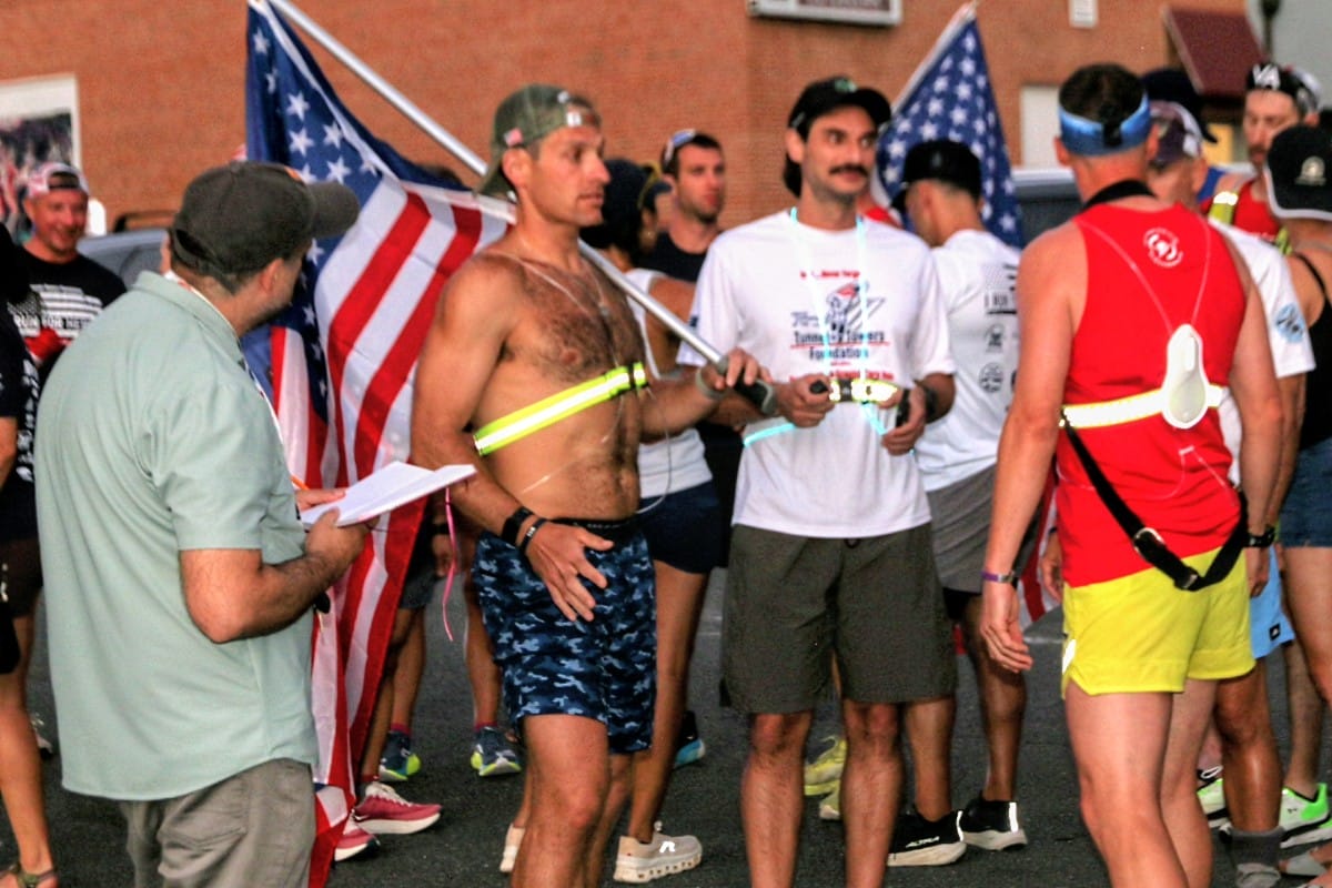 Runners gather at a race finish or award area, holding an American flag with medals visible in the crowd.
