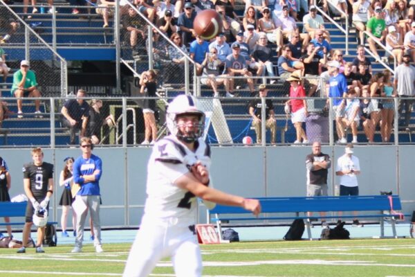 Quarterback in white jersey throws a pass from the field, with a crowded bleacher crowd behind him and sideline players nearby.