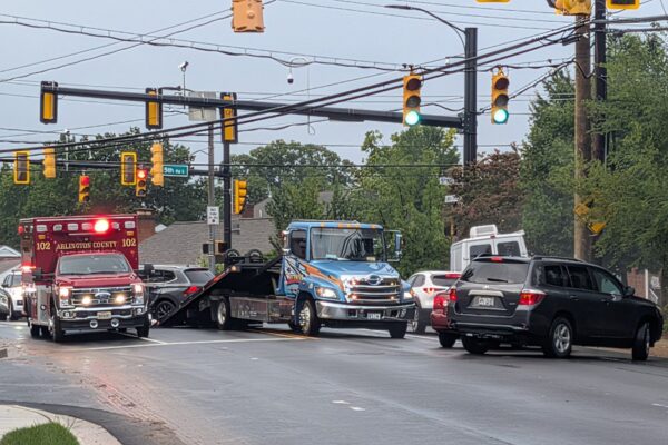 Emergency scene at an intersection: red Arlington County ambulance on the left, a blue tow truck lifting a damaged car, and surrounding vehicles nearby.
