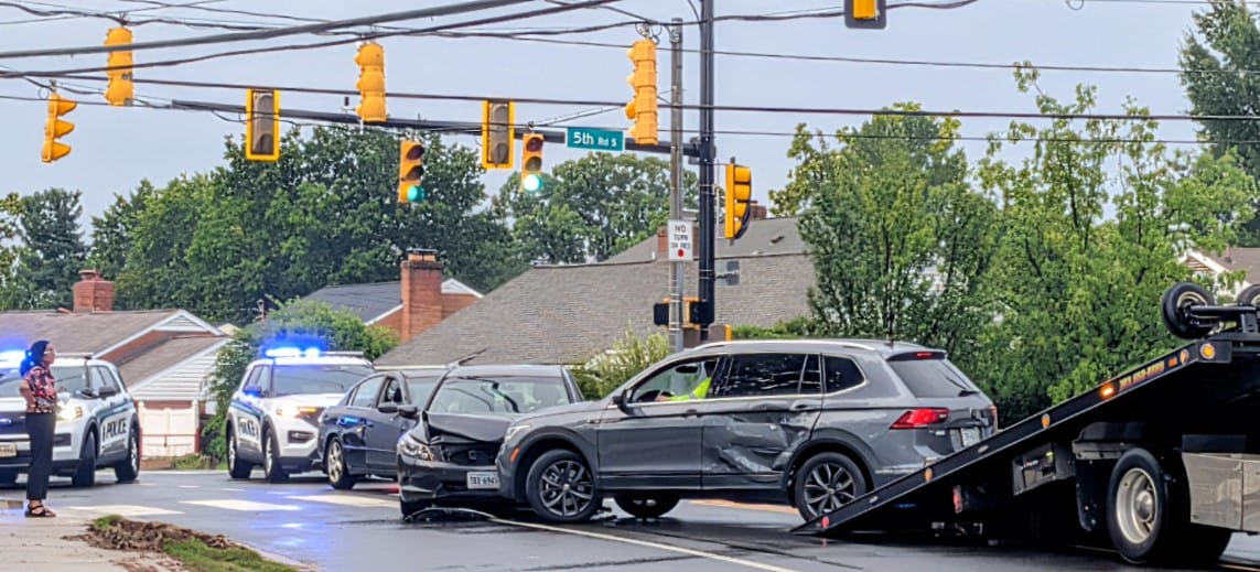 Gray SUV with major side damage sits on a tow truck at a busy intersection, police cars with flashing lights nearby, and bystanders watching the scene.