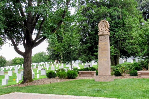 Grassy cemetery with rows of white headstones and a tall carved obelisk monument under trees.