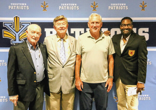 Four men pose for a group photo at a Yorktown Patriots event, standing in front of a blue backdrop with Patriots logos and text.