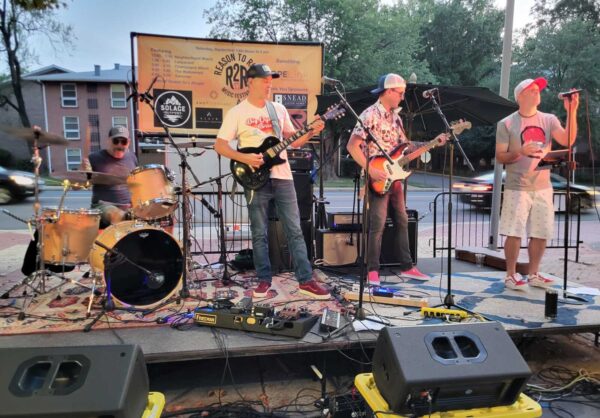 Outdoor street-side band performance with drummer, two guitarists, and a singer on a small stage with amplifiers and a banner behind them