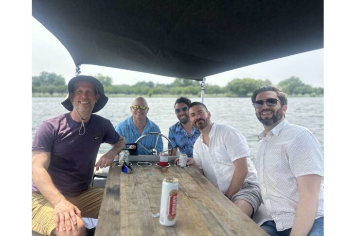 Five friends on a boat enjoying drinks under a canopy, smiling at the camera with a wooden table and water in the background.