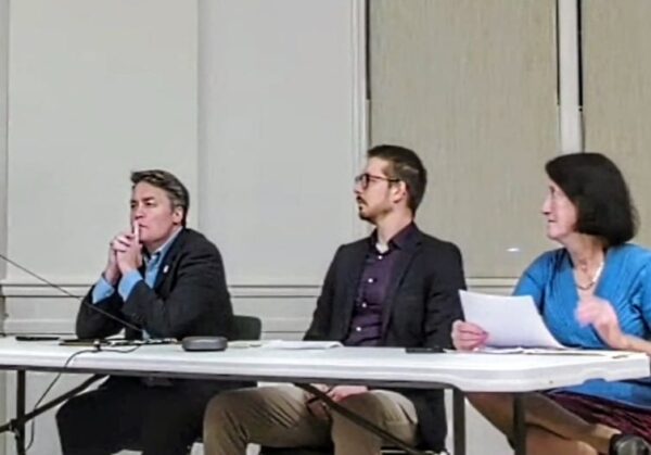 Three adults sit at a white panel table during a formal discussion: a man in a suit on the left, a man in a dark blazer in the middle, and a woman in blue on the right holding papers.