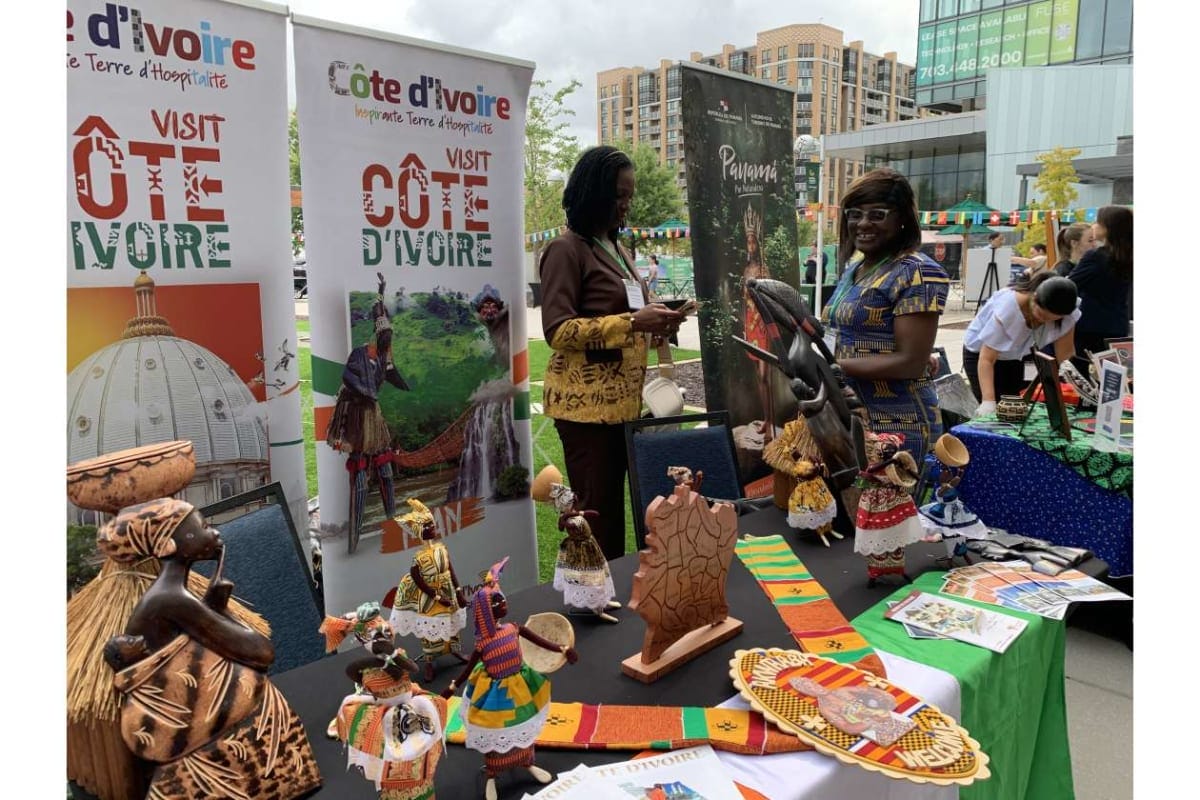 Outdoor market booth promoting Côte d'Ivoire with banners, displaying colorful handicrafts and dolls; two women chat nearby.