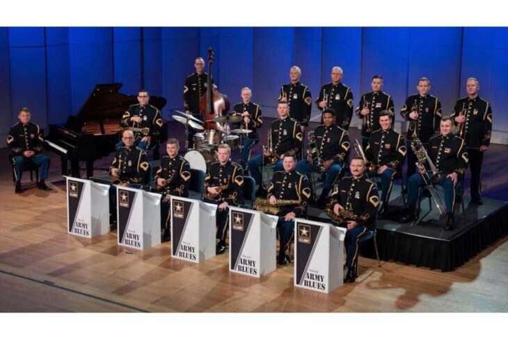U.S. Army Blues jazz ensemble in dress uniform on a blue-lit stage, about 20 musicians with brass and rhythm instruments, front row seated with 'Army Blues' music stands.