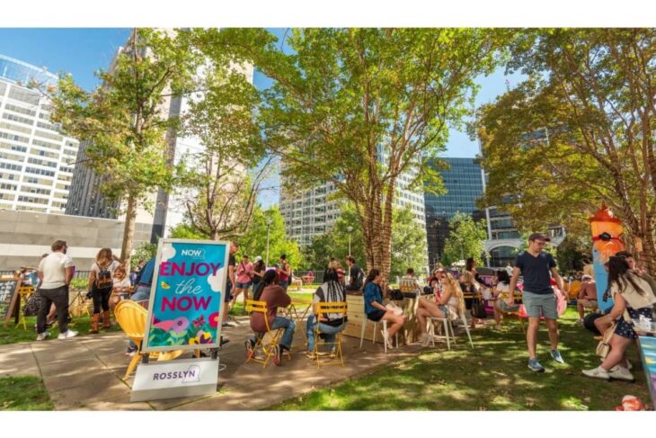 People socializing at a sunny outdoor event in a city park, seated on colorful chairs around tables near a sign that reads 'Enjoy the Now'.