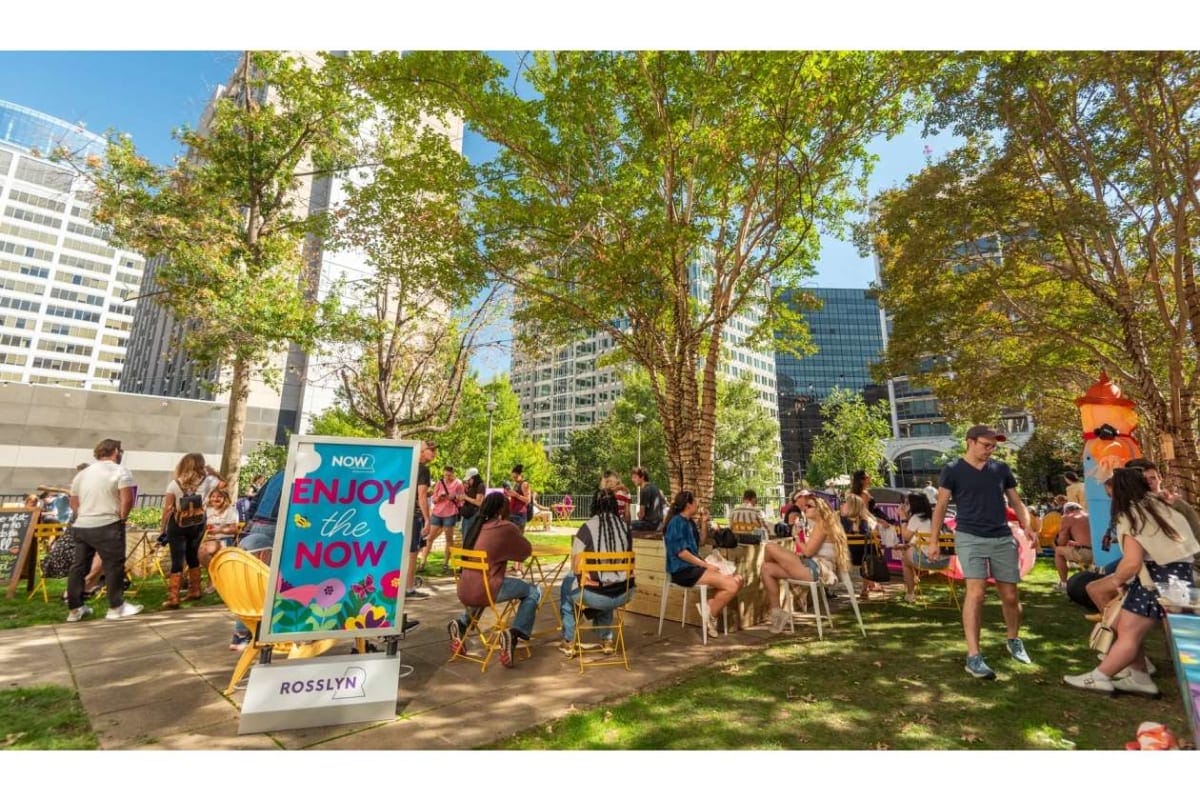 People socializing at a sunny outdoor event in a city park, seated on colorful chairs around tables near a sign that reads 'Enjoy the Now'.
