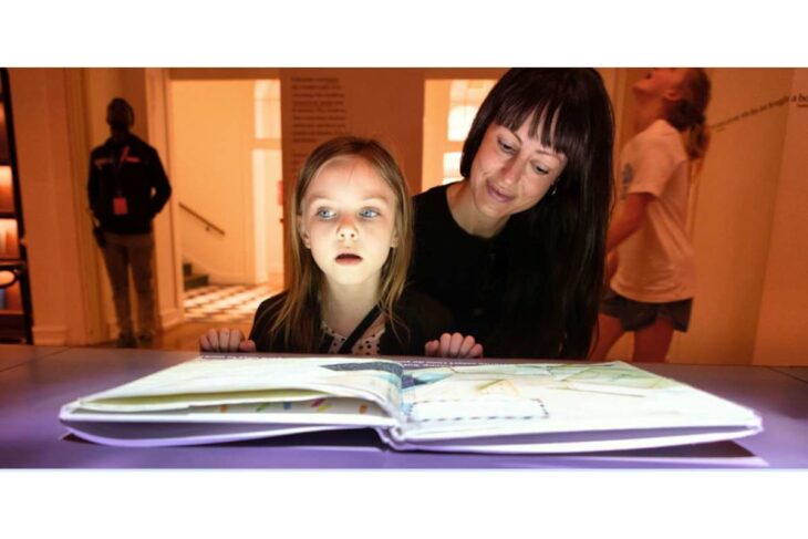 Mother and young girl lean over a glowing open book on a desk in a museum or exhibit space, reading together.