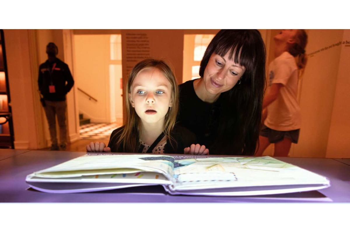 Mother and young girl lean over a glowing open book on a desk in a museum or exhibit space, reading together.
