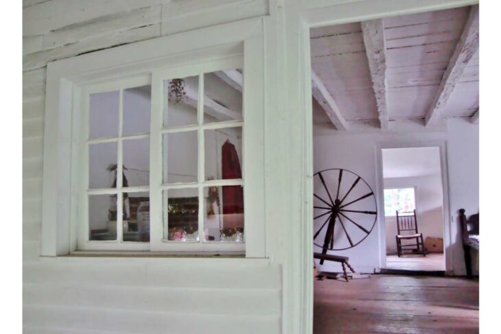 White-washed interior with a pane-window inset, a large black wagon wheel leaning against a wall, and wooden beams overhead in a rustic room.