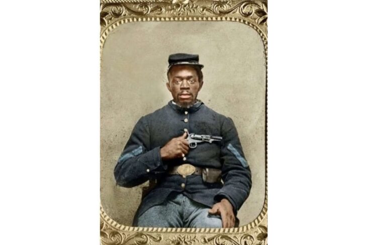 Civil War–era portrait of an African American man in a dark blue military uniform, seated, holding a pistol across his chest with a belt and small cap.