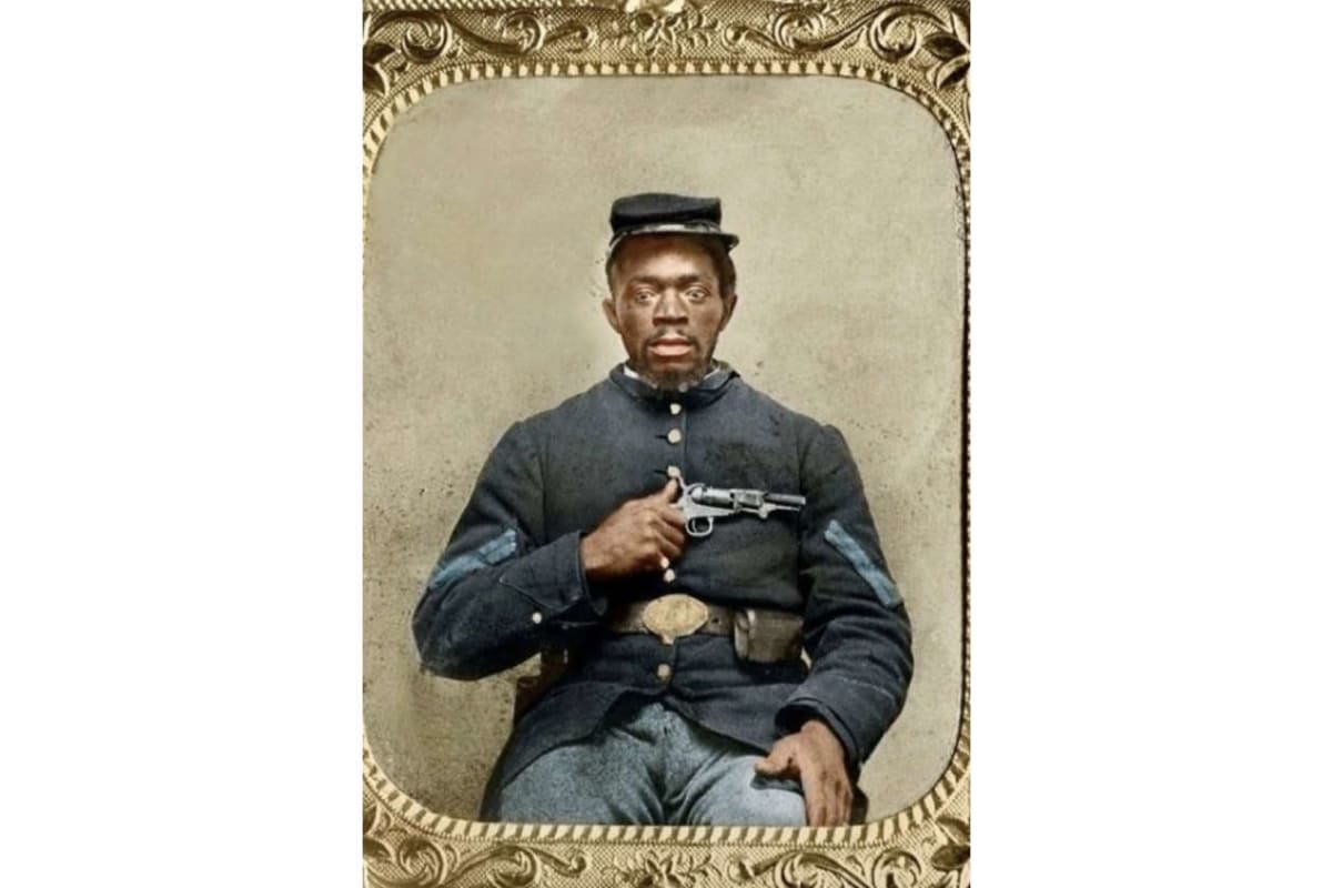 Civil War–era portrait of an African American man in a dark blue military uniform, seated, holding a pistol across his chest with a belt and small cap.