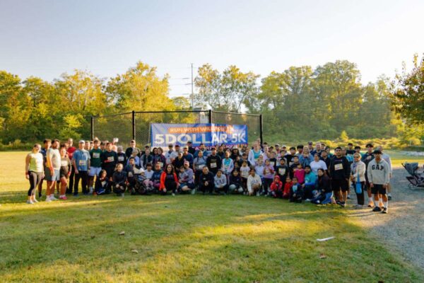 Wide shot of a large group of runners and supporters posing in a park with a banner reading '5 Dollar 5K' behind them.