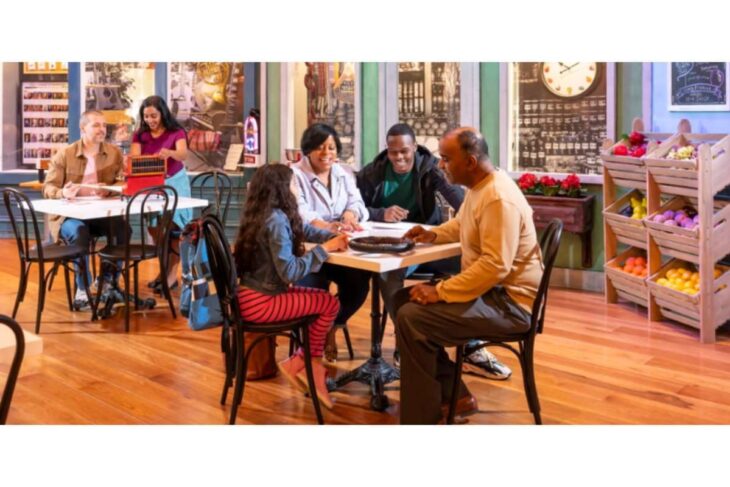 A diverse group of people sit around a table in a bright cafe, chatting and sharing a meal; fruit stands visible in the background right side.