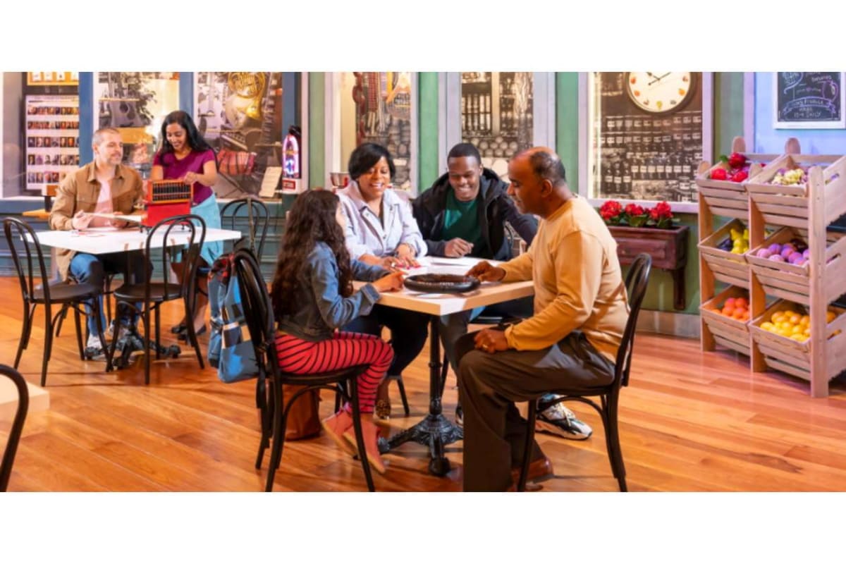 A diverse group of people sit around a table in a bright cafe, chatting and sharing a meal; fruit stands visible in the background right side.