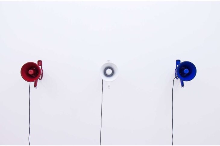 Three wall-mounted circular speakers in red, white, and blue with cords hanging down on a white wall.