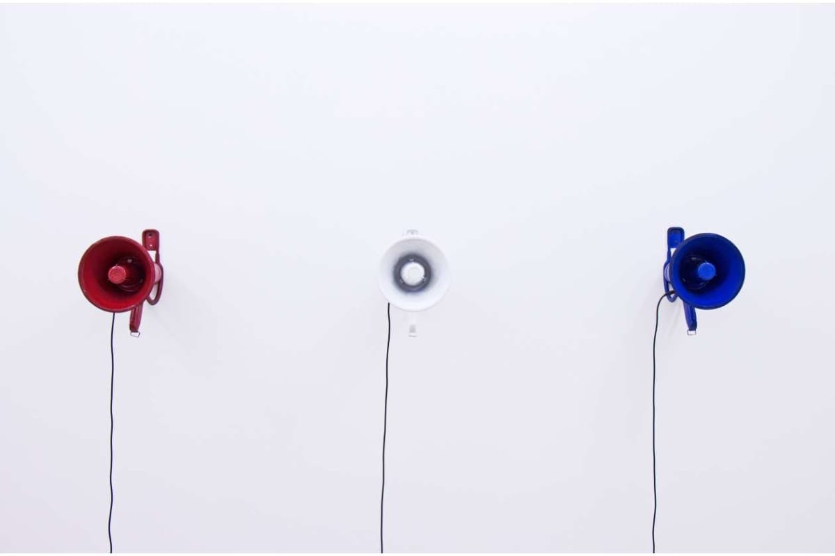 Three wall-mounted circular speakers in red, white, and blue with cords hanging down on a white wall.