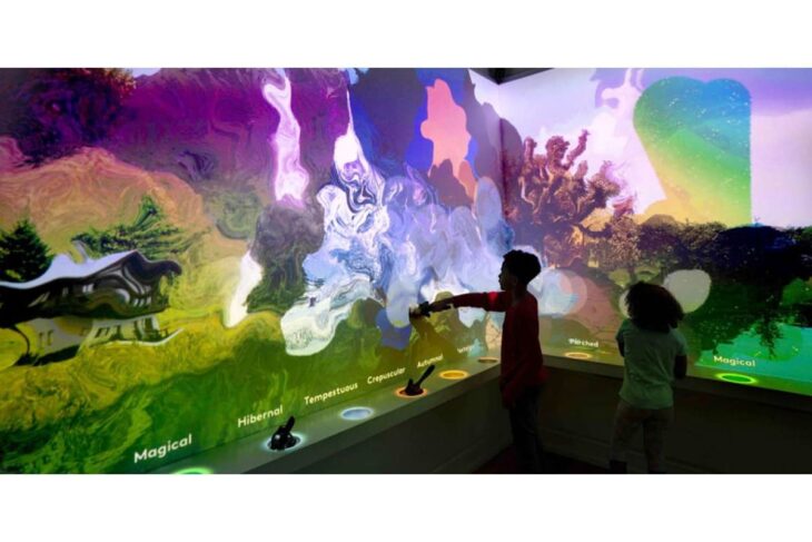Two children interact with a vivid, multicolored wall exhibit featuring swirling patterns and glowing labeled sections at the bottom.