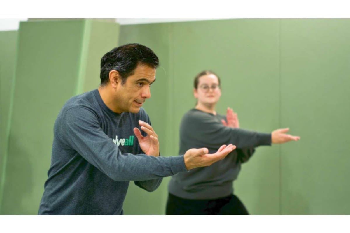 Two adults practicing martial arts in a gym; the man in the foreground demonstrates a hand technique while a woman follows in the background, both extending their arms.