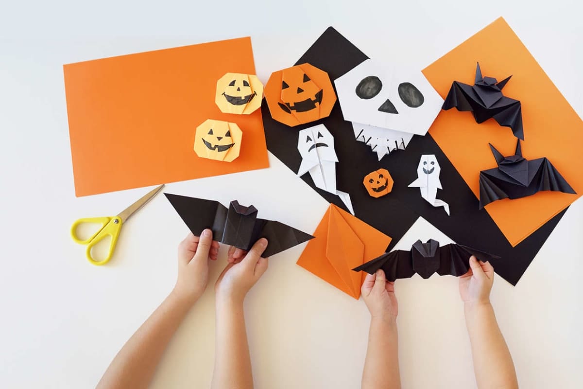 Hands crafting origami bats from black paper among orange pumpkins and ghost shapes on orange/black sheets, scissors nearby