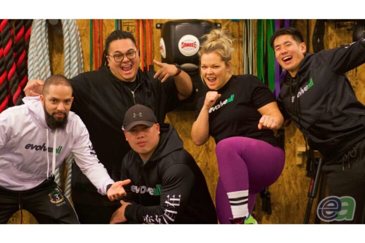 Group of five diverse fitness enthusiasts posing energetically in a gym, wearing EvolvAll branded apparel with ropes and gear in the background.