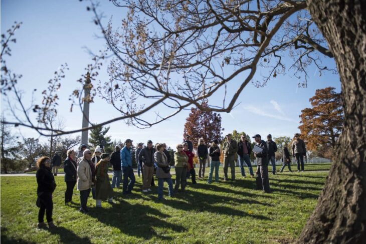 Group of people outdoors in a park listening to a guide with a clipboard, near an obelisk and autumn trees.