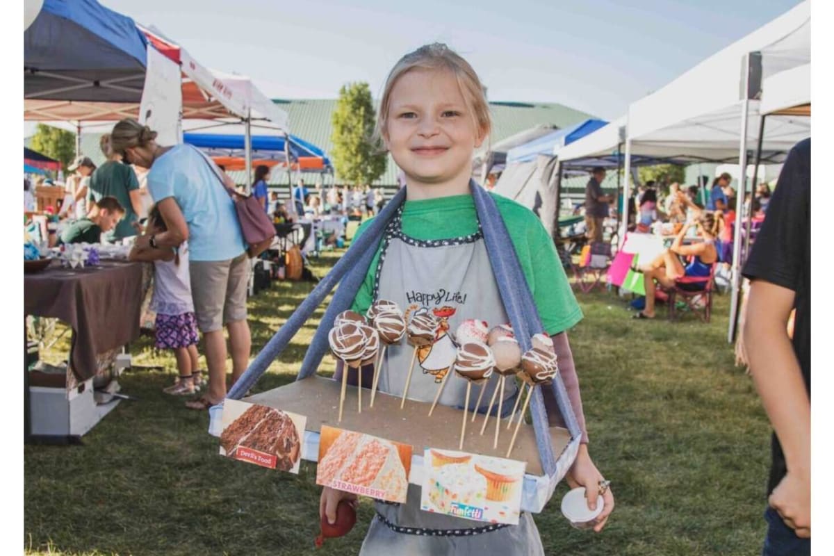 Smiling girl at an outdoor market, selling chocolate‑drizzled candy apples on sticks from a box.