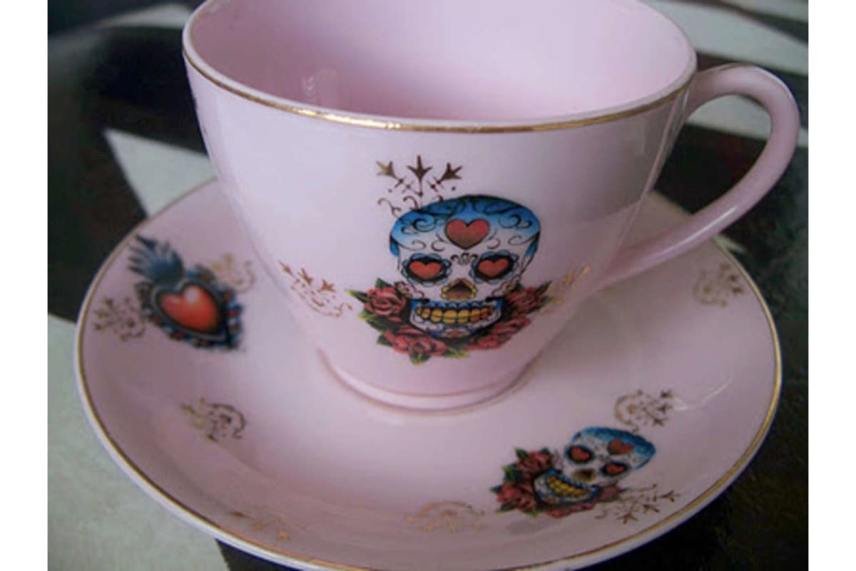 Pink teacup with a matching saucer, both pale pink with gold trim and sugar skull heart motifs.