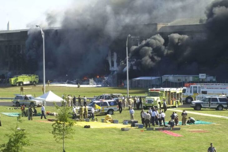 Large building on fire with thick black smoke; firefighters and emergency vehicles on scene in a grassy area outside the structure.