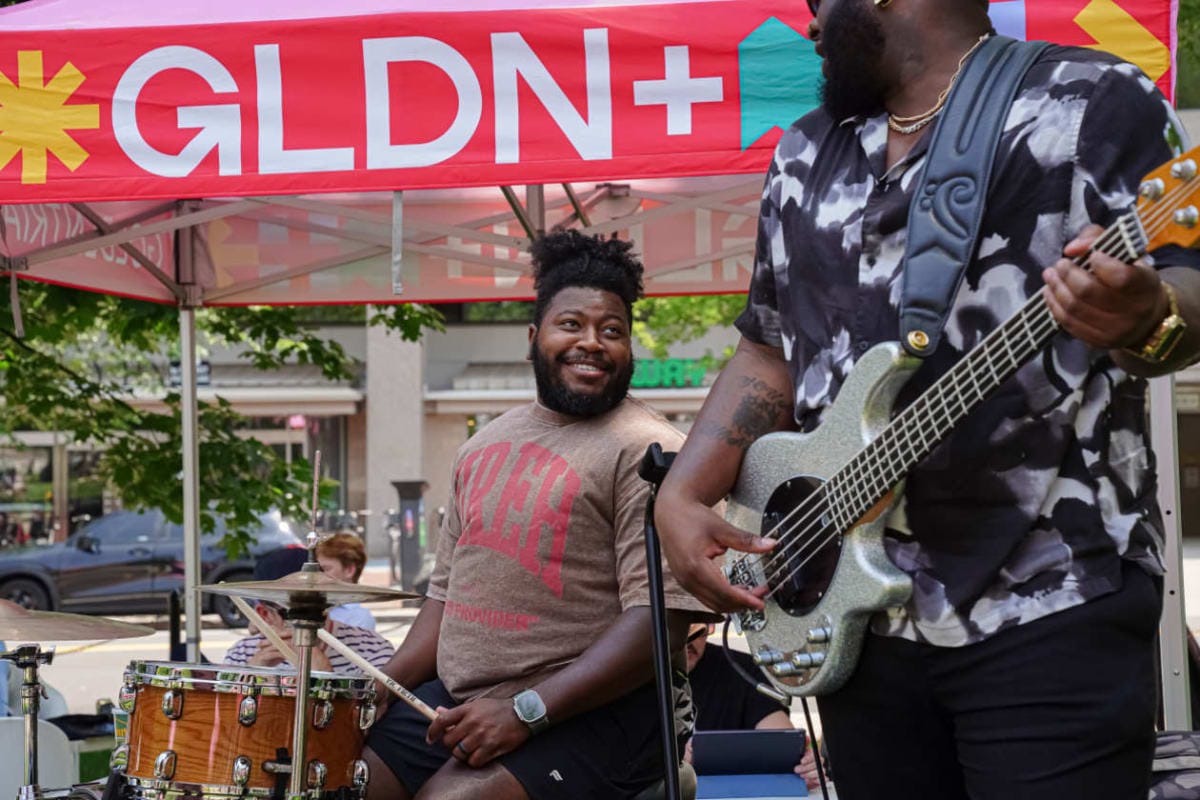 Two male musicians performing outdoors under a red GLDN+ banner; one on drums smiling, wearing a brown shirt and watch, the other playing a silver bass guitar.