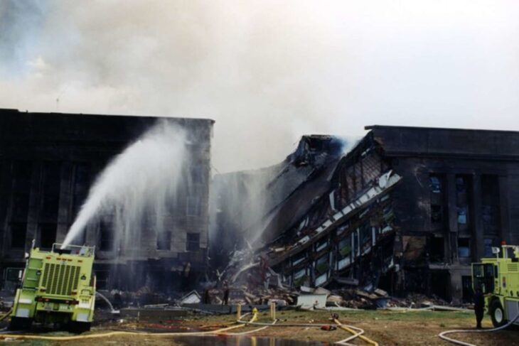 Firefighters spray water on a partially collapsed building with a tilted roof and debris on the ground, two yellow fire engines nearby.